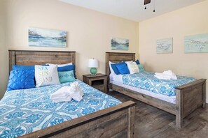 Une chambre d’hôtel avec deux lits en bois, chacun recouvert d’une couvre-lit à motifs bleus et agrémenté d’oreillers assortis. Des tableaux sont accrochés au mur au-dessus des lits, et une petite table de chevet avec une lampe se trouve à côté de chaque lit.