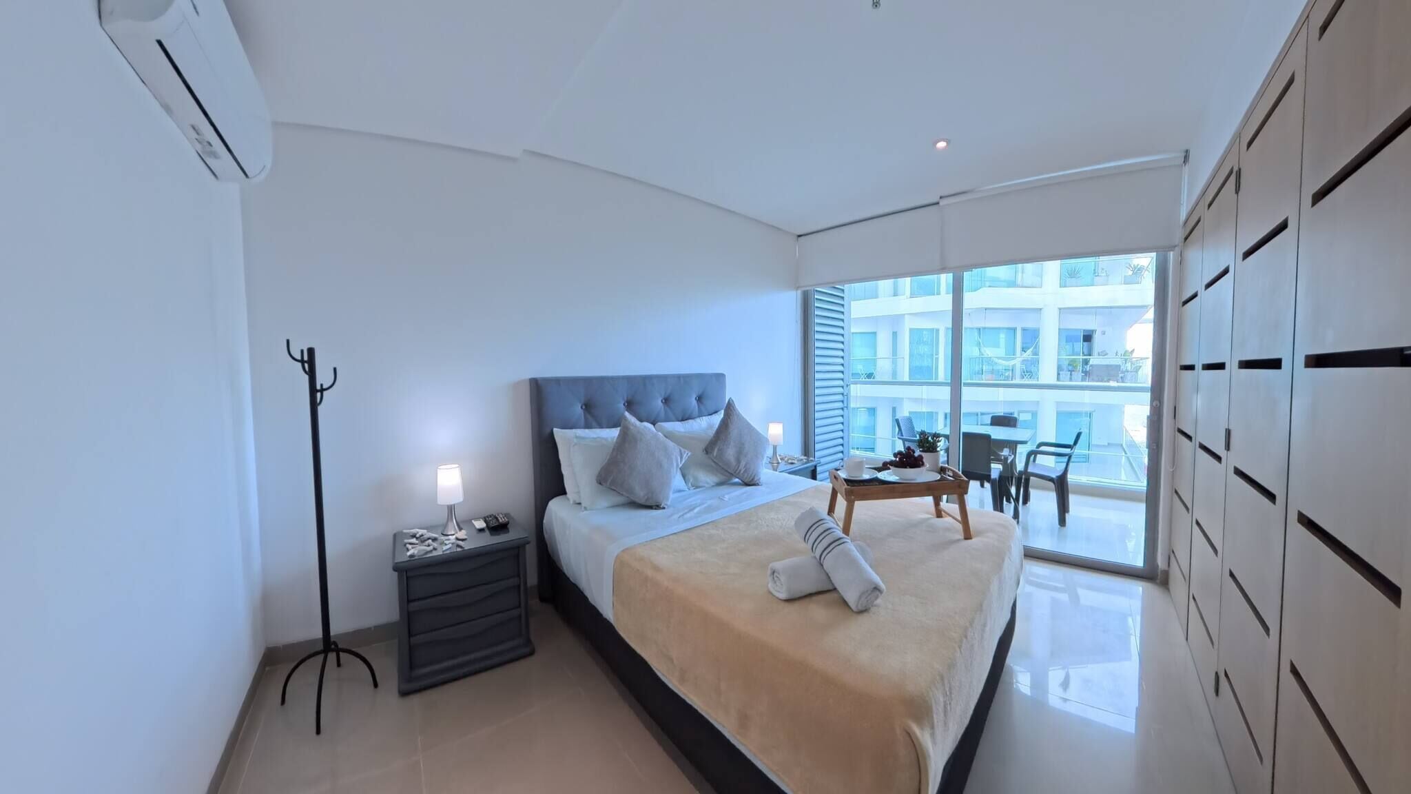 Une chambre d’hôtel moderne avec un grand lit, une table de chevet avec une lampe, un fauteuil et une vue sur la ville grâce à de grandes fenêtres.