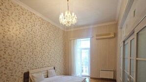 Un dormitorio con piso de madera, una cama con ropa blanca, una araña y un ventanal con cortinas.