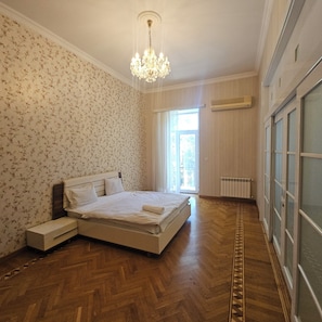 Un dormitorio con piso de madera, una cama con ropa blanca, una araña y un ventanal con cortinas.