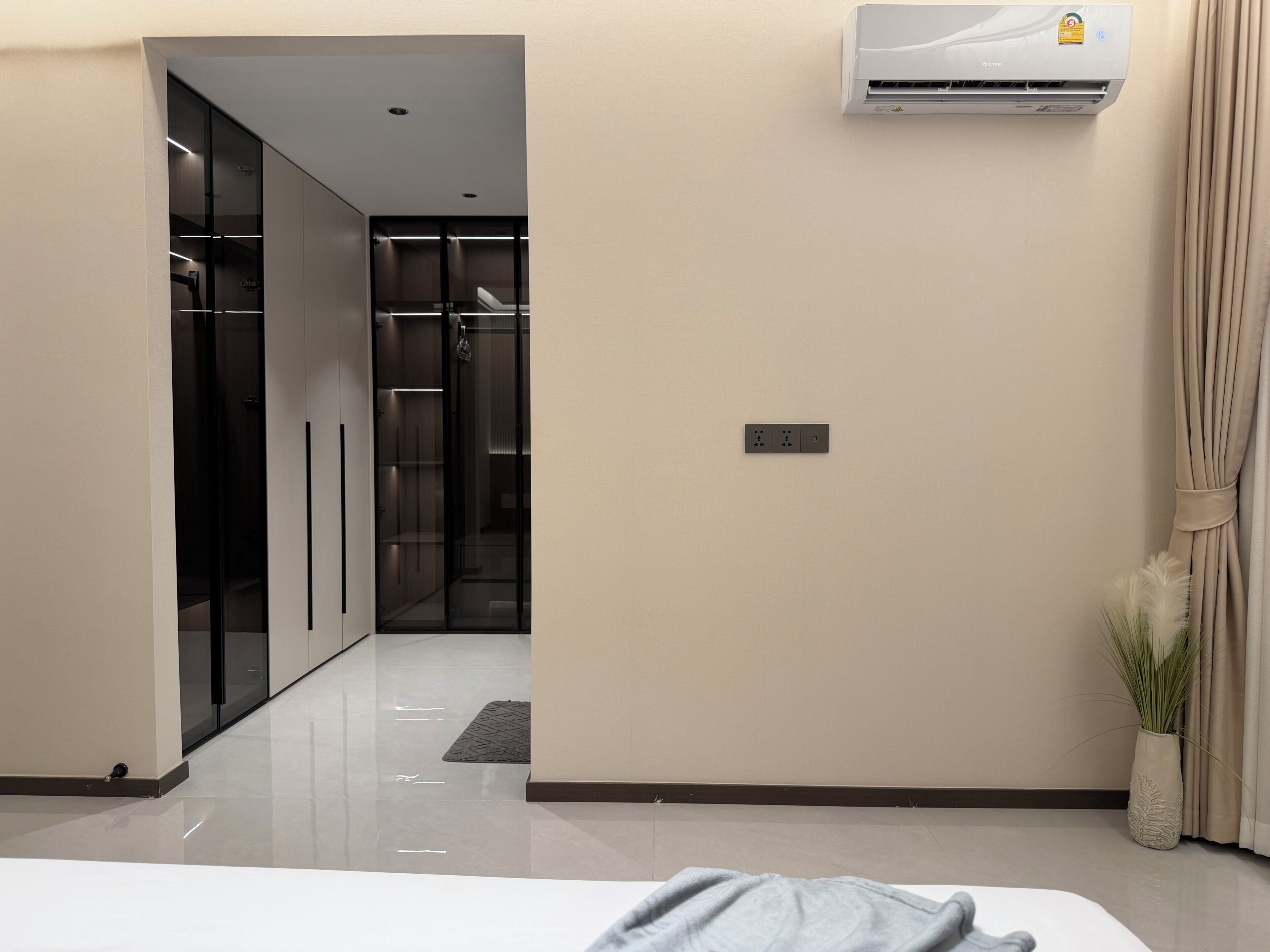 A hotel room with a bed, an air conditioning unit, a power outlet, and a potted plant.