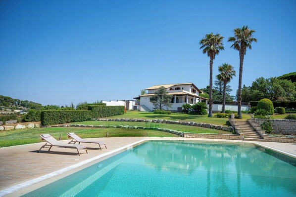 Une piscine avec des chaises longues, une maison avec un toit en tuiles et des palmiers.