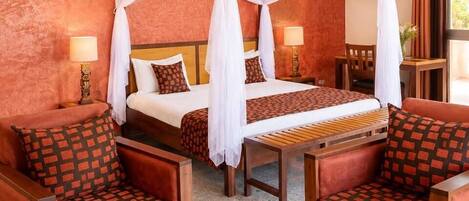 Habitación de hotel con cama con dosel, dos sillones, una mesita con un jarrón de flores y una mesita de noche con lámpara.