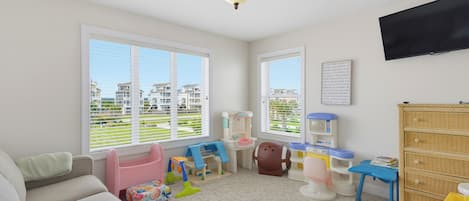 A well-lit room with a large window, a ceiling fan, a television, and a crib.