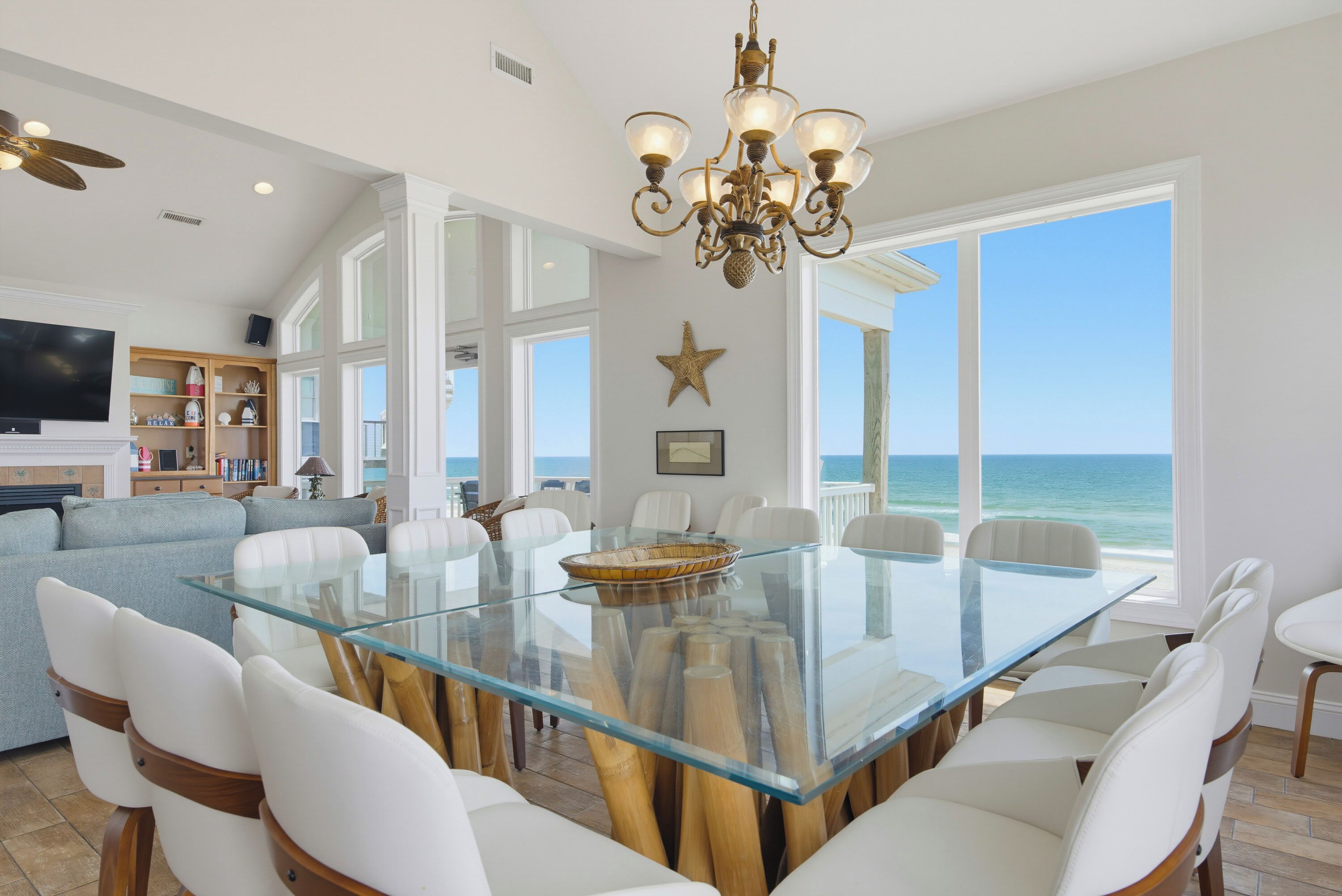 A spacious dining area with a glass-top table surrounded by white chairs, a chandelier, and a view of the ocean.