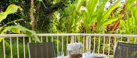 An outdoor dining area with a white table set for four, surrounded by chairs, with a view of lush greenery.