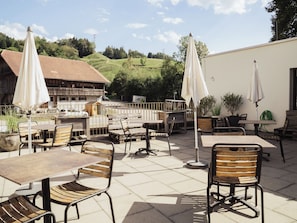 Un patio al aire libre con mesas, sillas, sombrillas y vista a un edificio y colinas al fondo.