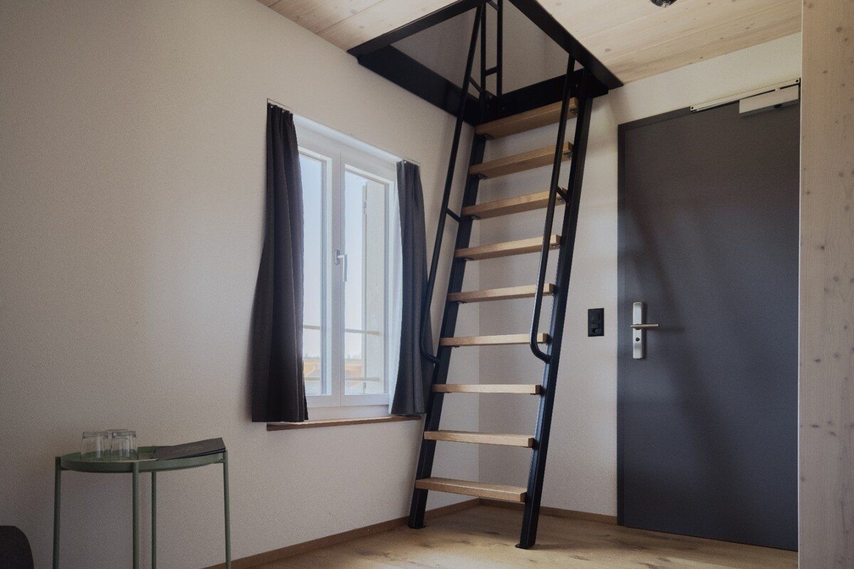 Una habitación con una escalera de madera, una ventana con cortinas, una puerta y una pequeña mesa con vasos.