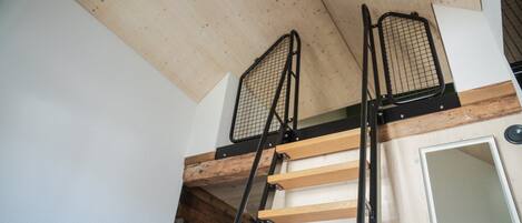 A loft-style room with a wooden ceiling, a metal ladder, and a door leading to another space.