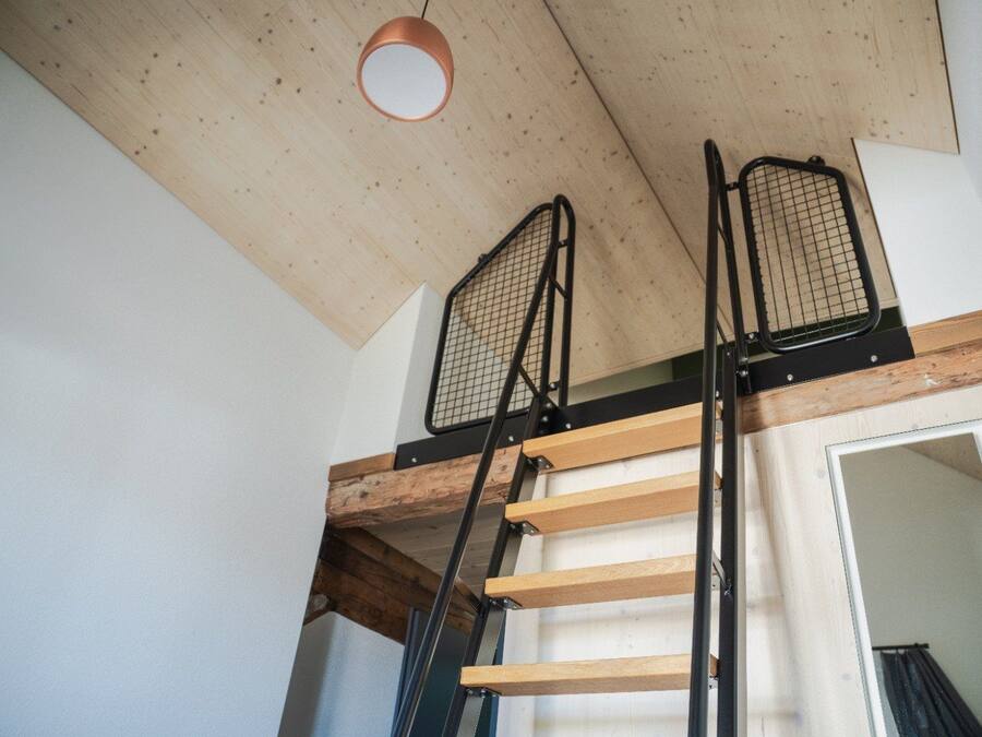 A loft-style room with a wooden ceiling, a metal ladder, and a door leading to another space.