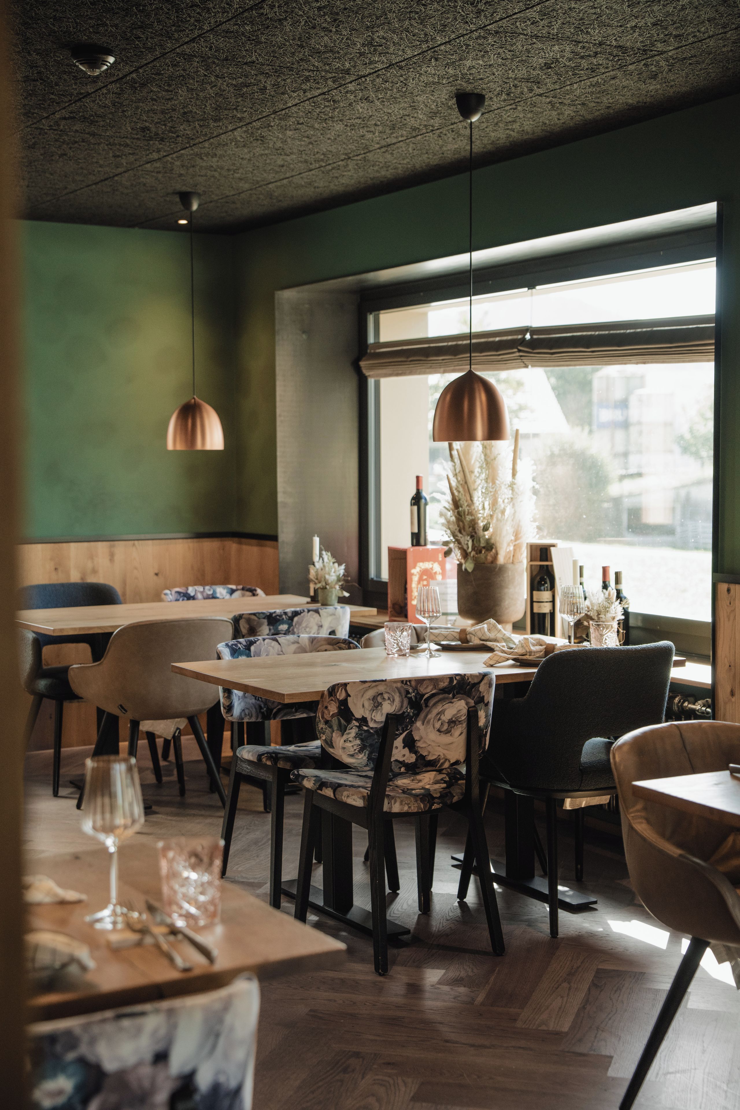 A restaurant interior with wooden tables, chairs, hanging pendant lights, and a large window providing natural light.