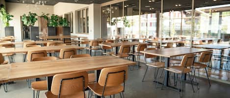 Un amplio comedor interior con múltiples mesas y sillas de madera, grandes ventanales que dejan entrar luz natural, y plantas en macetas.