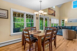 A dining area with a wooden table and chairs, large windows with a view of greenery, and a ceiling light fixture. - 5BR Home Quietside of MDI - Sleeps 23! [Headlands] (Tremont)