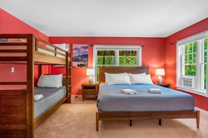 Ein Schlafzimmer mit Etagenbett, Doppelbett, zwei Nachttischen mit Lampen, einem Fenster mit Blick auf Grünflächen und einem Bild an der Wand.