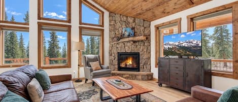 A cozy living room with a stone fireplace, leather sofas, a television, and large windows offering a view of trees.