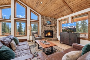 A cozy living room with a stone fireplace, leather sofas, a television, and large windows offering a view of trees. - Cozy Fairplay Log Cabin | Arcade & Mountain Views (Fairplay)