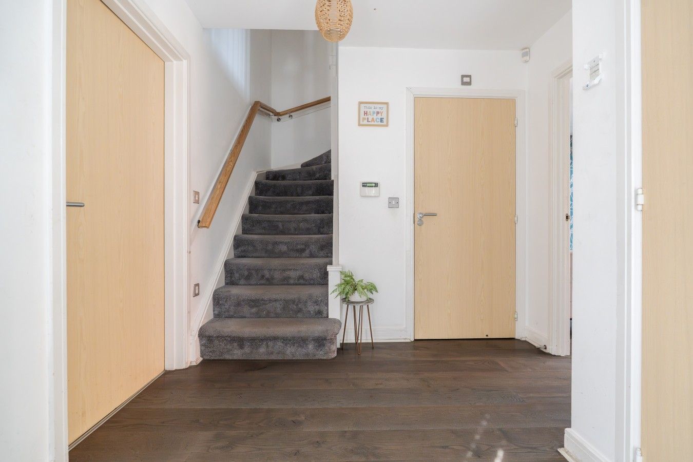 Una escalera con barandilla que conduce a una puerta, una planta pequeña sobre una mesita auxiliar y un aplique de pared.