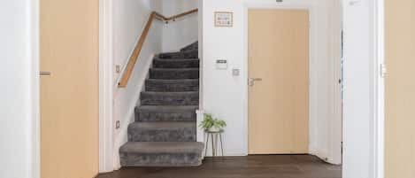 Una escalera con barandilla que conduce a una puerta, una planta pequeña sobre una mesita auxiliar y un aplique de pared.