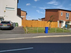 Un carrer residencial amb cases, un cotxe estacionat i un cubell de brossa blau.