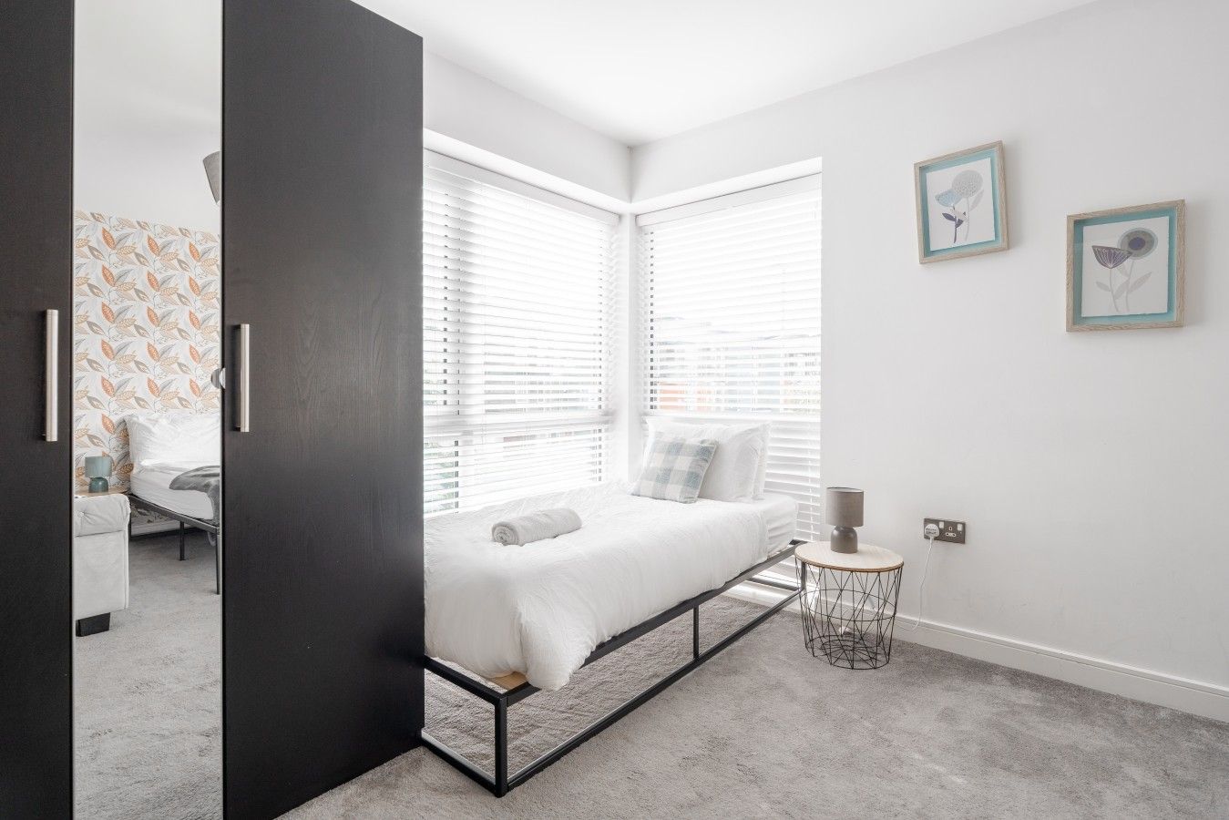 A modern bedroom with a bed, two windows, a bedside table, and a wall with two framed pictures.