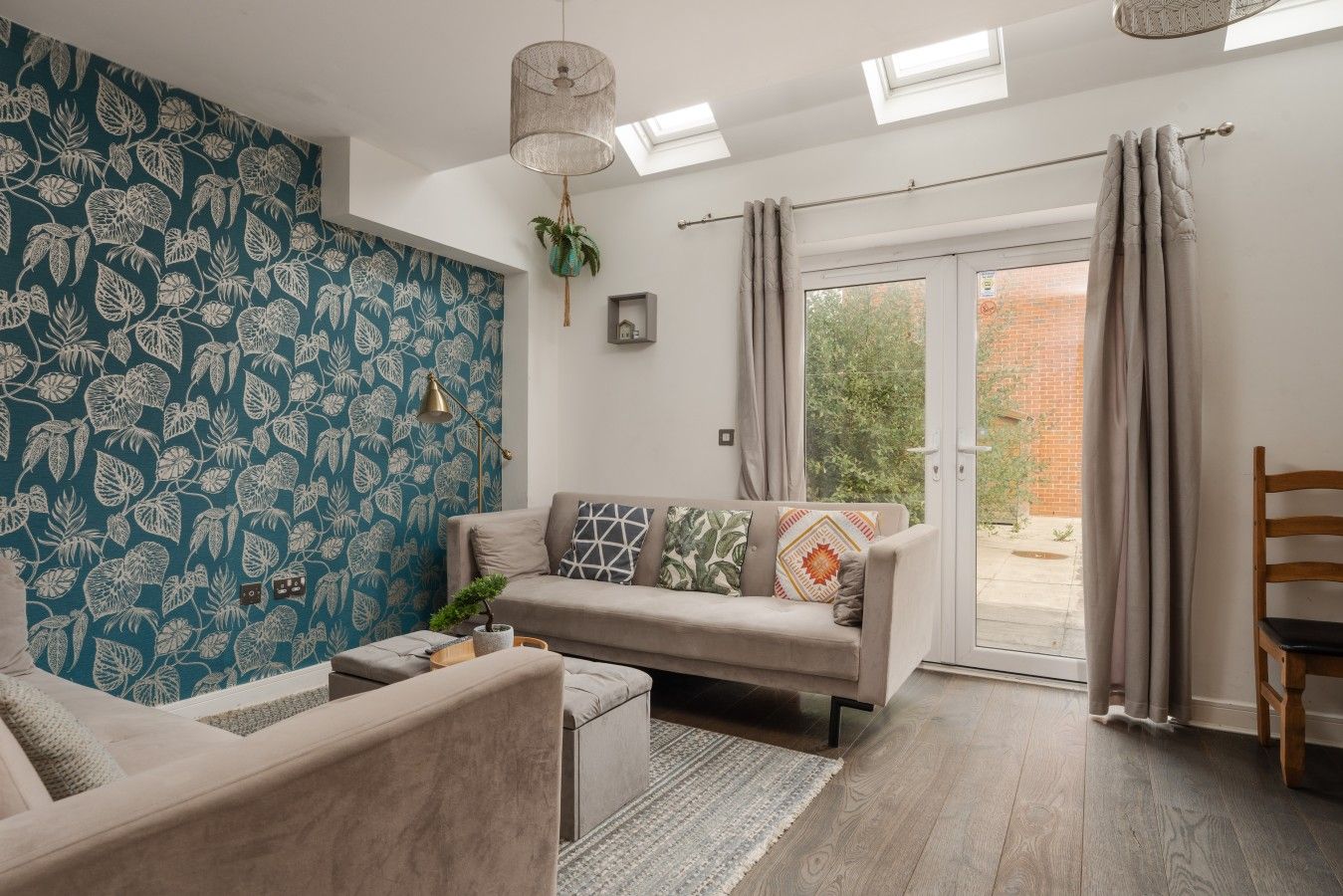 A living room with a sofa, a coffee table, and a patterned wall. There is a window with curtains and a view of greenery outside.