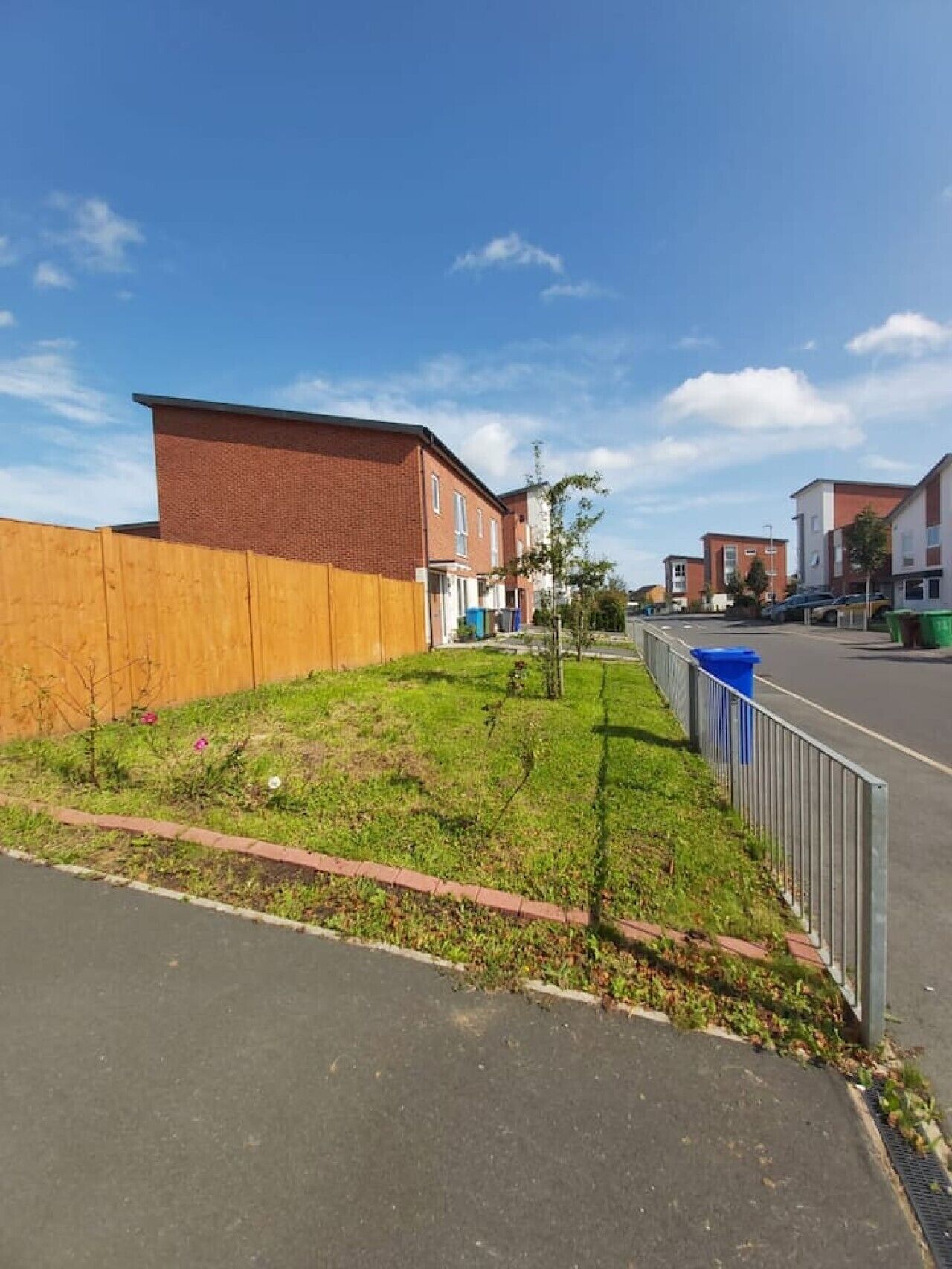A residential street with houses, a grassy area, and a fence.