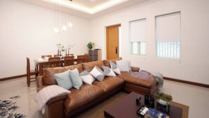 A modern living room with a brown leather sofa, dining table, and chairs. There is a wooden door, two windows, and a ceiling light fixture. - Spacious Heritage House Prime Location Family Stay (Bangkok)