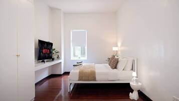 A bedroom with a bed, a TV on a shelf, a window, and a bedside table with a lamp.