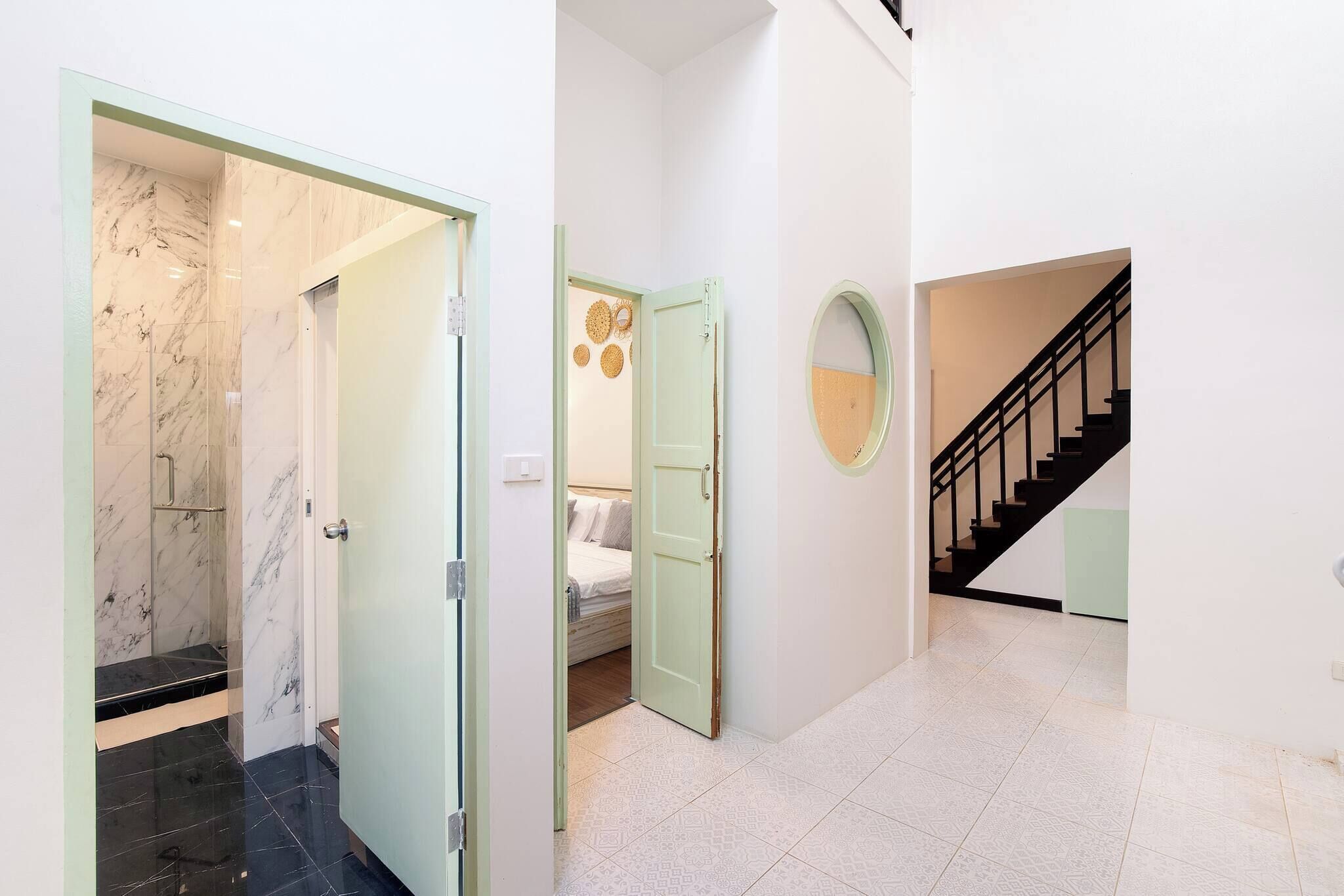 Un couloir avec une porte en verre donnant sur une salle de bain, une chambre à coucher avec un lit et un miroir fixé au mur, et un escalier avec une rampe noire.