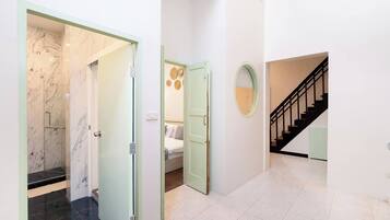A hallway with a glass door leading to a bathroom, a bedroom with a bed and a wall-mounted mirror, and a staircase with a black railing.
