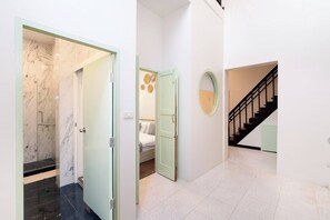 Un couloir avec une porte en verre donnant sur une salle de bain, une chambre à coucher avec un lit et un miroir fixé au mur, et un escalier avec une rampe noire.