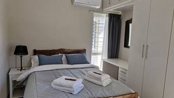 A bedroom with a bed, bedside table, lamp, air conditioning unit, window with curtains, and a wardrobe.