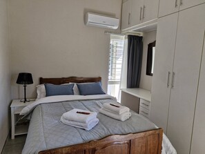 A bedroom with a bed, bedside table, lamp, air conditioning unit, window with curtains, and a wardrobe.