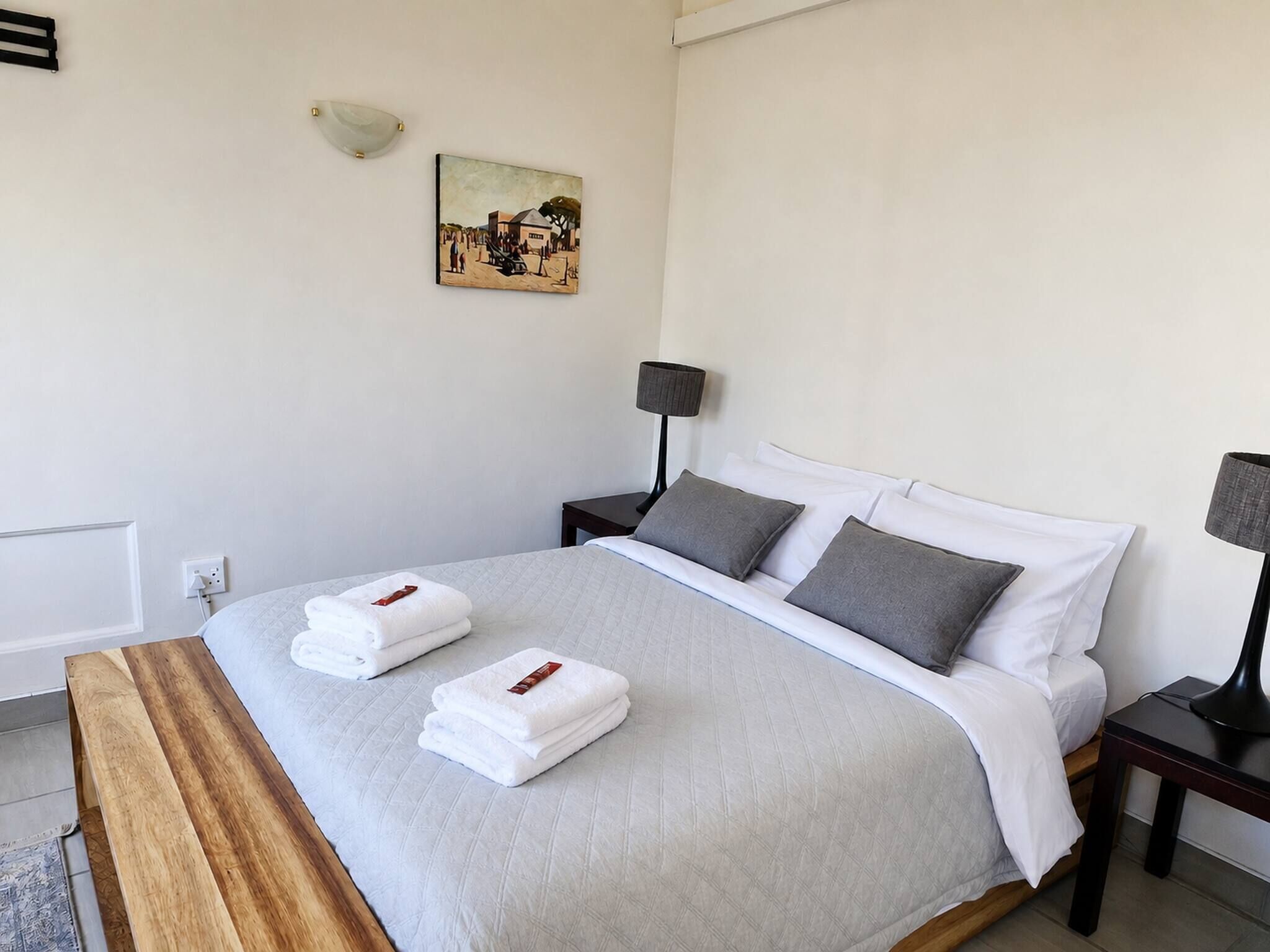 A neatly made bed with white linens and two pillows, a bedside table with a lamp, and a painting on the wall.