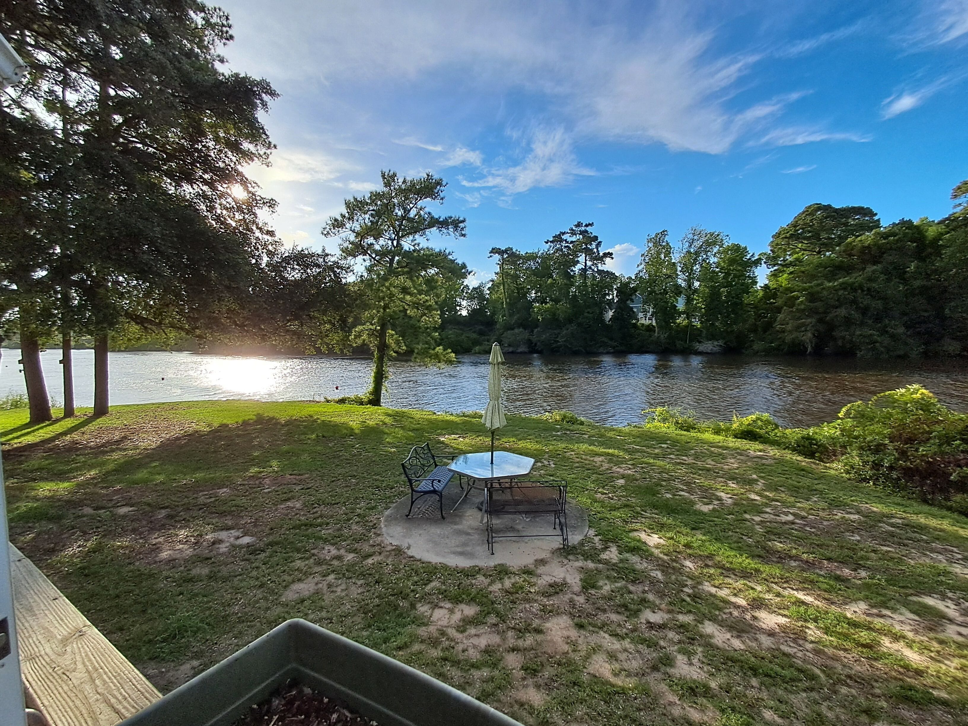 A lakeside patio with a table and chairs, surrounded by trees and a view of the lake.