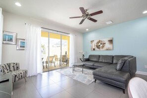 A living room with a grey sofa, a coffee table, a ceiling fan, and a sliding glass door leading to an outdoor patio. - Charming 3 BR with pool at Serenity (Clermont)