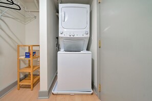 A compact laundry room with a stacked washer and dryer, a wooden shelf with laundry products, and a metal drying rack. - Chic City Loft - Nearby Parking - Hospitals - 2F (Louisville)