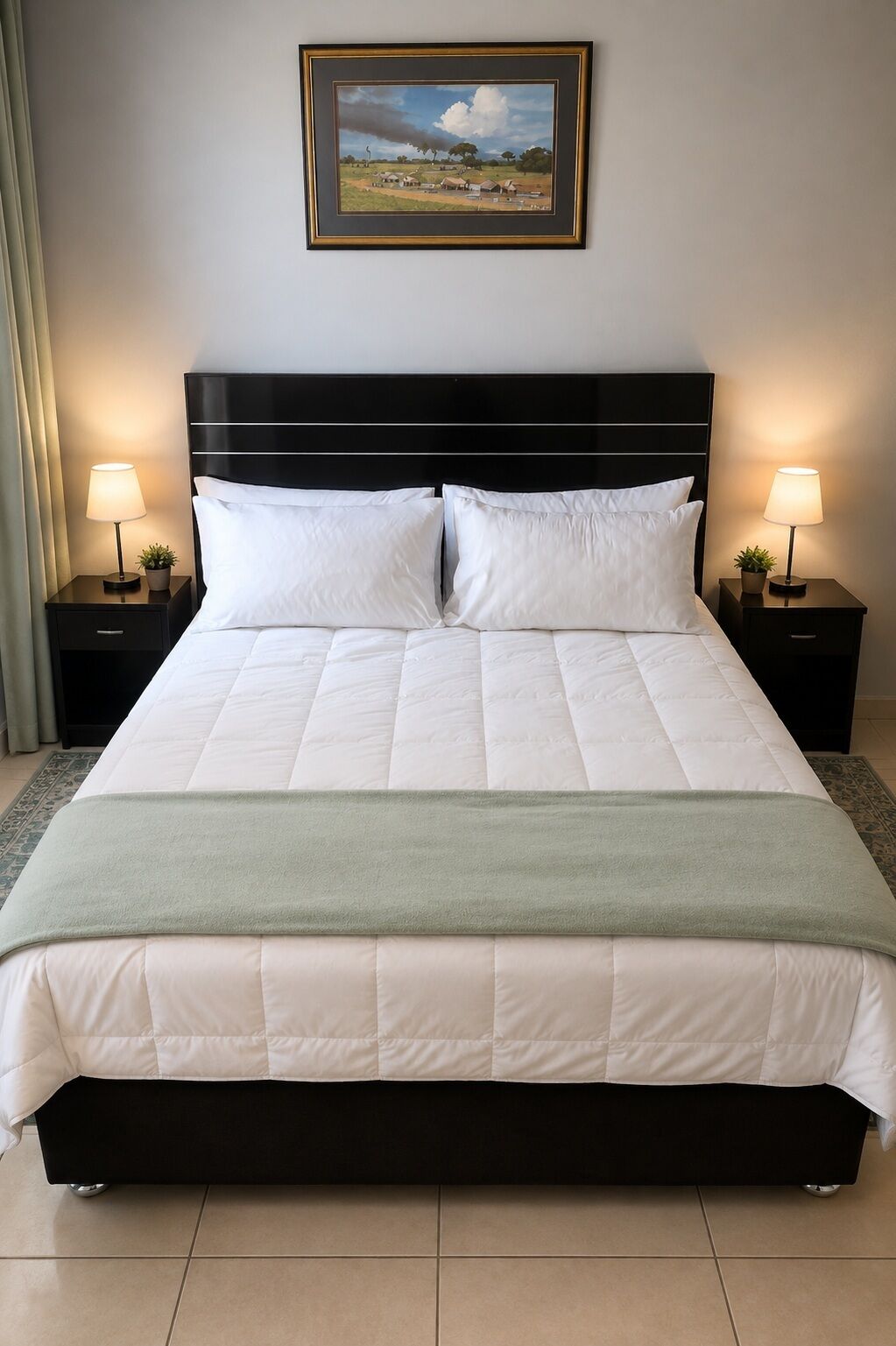 A neatly made bed with a quilted white duvet, two pillows, and a green throw blanket. There are two bedside tables with lamps and a framed landscape painting above the bed.