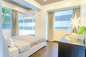 Quarto de hotel moderno com uma cama grande, uma escrivaninha com luminária e janelas amplas com cortinas.