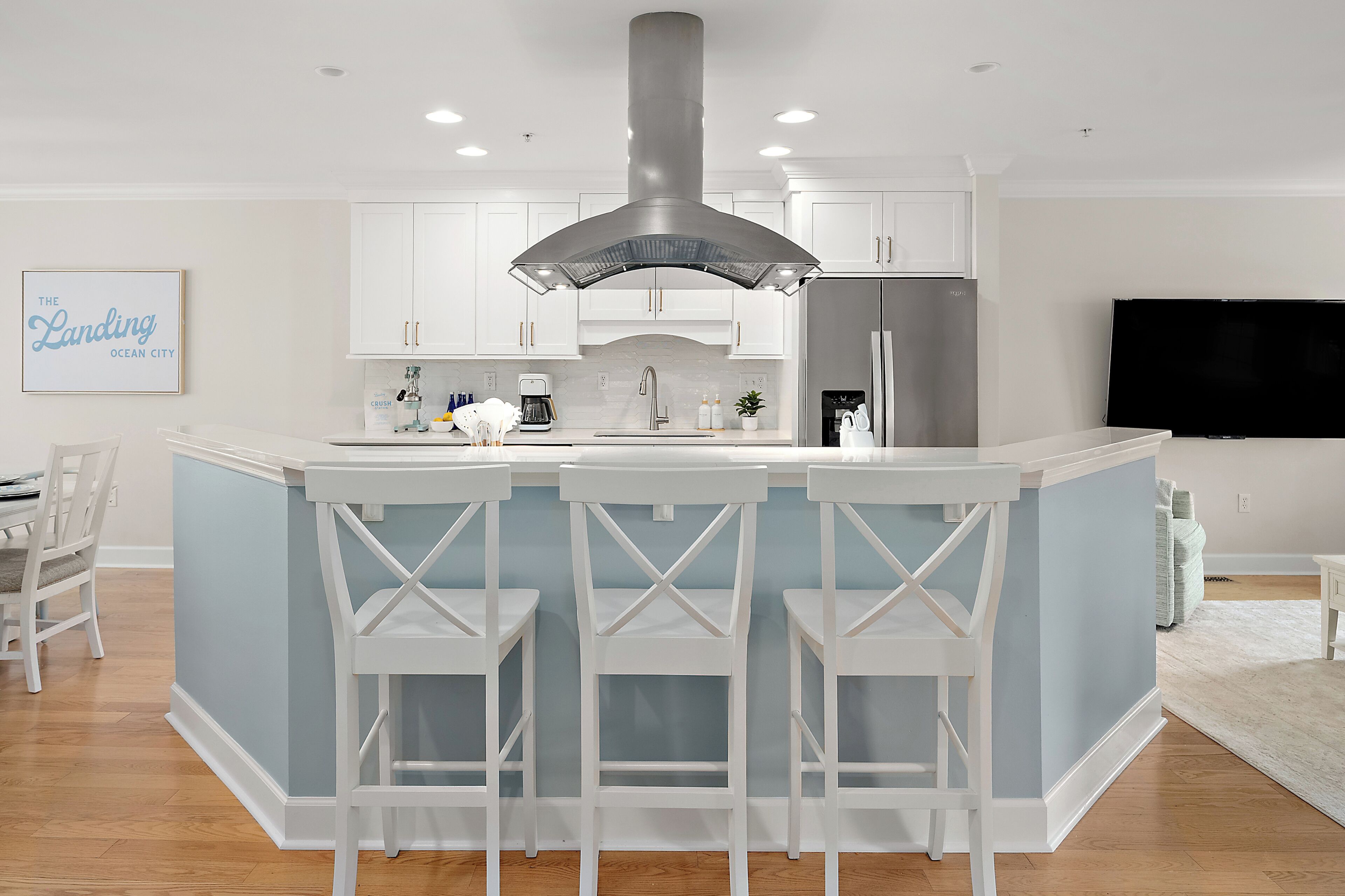 Una cocina moderna con muebles blancos, una isla central con taburetes de bar, un refrigerador de acero inoxidable y un televisor de pantalla plana montado en la pared.