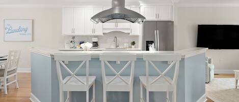 A modern kitchen with white cabinetry, a central island with bar stools, a stainless steel refrigerator, and a flat-screen TV mounted on the wall.
