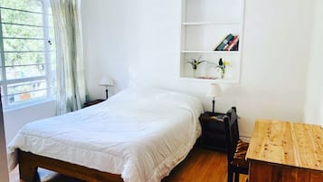 A bedroom with a wooden bed, a bedside table with a lamp, a small wooden table, and a white wall with a shelf holding books and a vase.