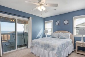 Ein Schlafzimmer mit einem Bett, einem Deckenventilator, einem Fenster mit Jalousien und Blick auf den Strand durch eine Schiebetür.