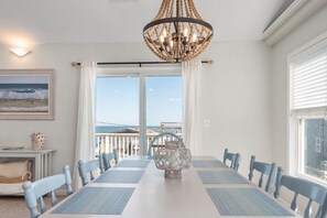Ein Esszimmer mit großem Tisch, Stühlen, einer Kristallleuchtre und Meerblick durch hohe Fenster.