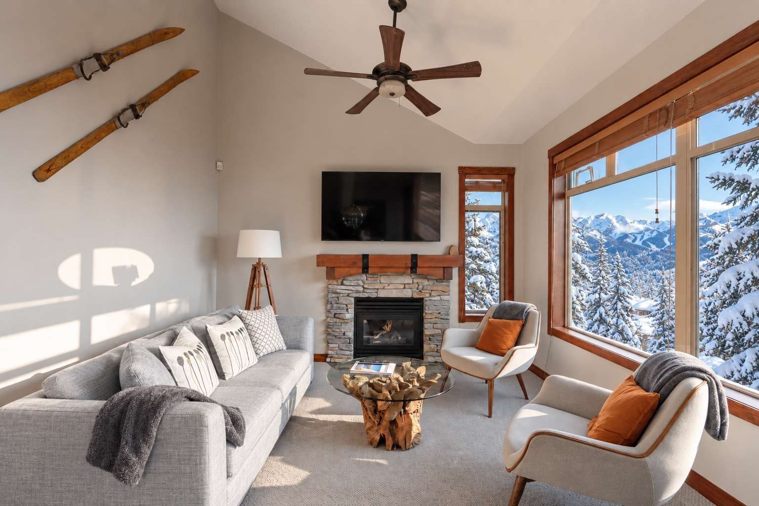 A living room with a stone fireplace, a ceiling fan, a large window with a snowy mountain view, and a gray sofa with multiple pillows.