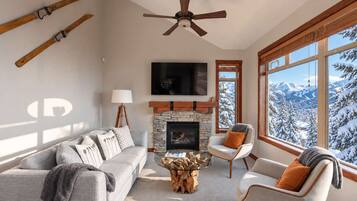 A living room with a stone fireplace, a ceiling fan, a large window with a snowy mountain view, and a gray sofa with multiple pillows.
