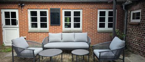 A brick house with a tiled roof, a white door, and a patio with a grey sofa and two chairs.