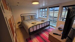 Una cama individual con estructura metálica, una mesita de noche, una silla, una pequeña mesa redonda y una ventana con vistas al exterior.