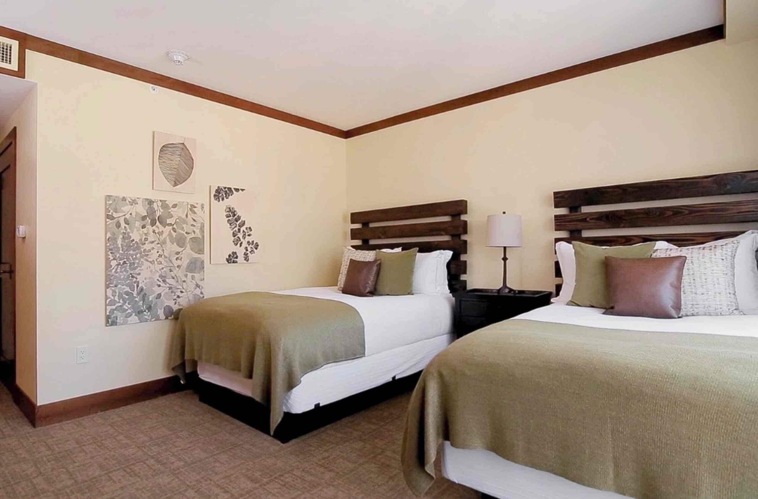 A hotel room with two beds, a nightstand with a lamp, and artwork on the walls.