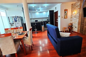 Sala de estar moderna com sofá azul, mesa de jantar de madeira e cadeiras. Uma cozinha visível ao fundo, com armários brancos e bancada preta.